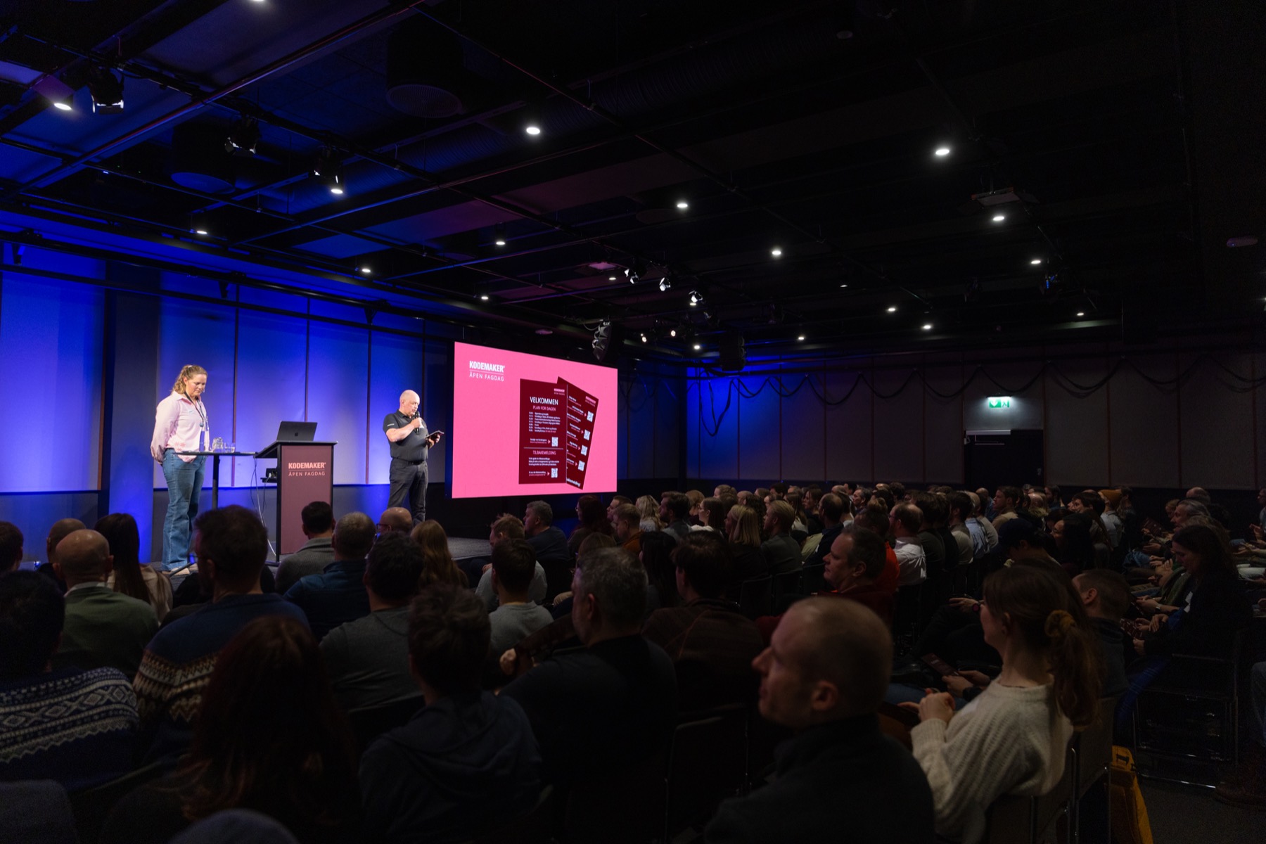 Fullsatt sal under Kodemaker Åpen Fagdag 2026, to personer på scenen foran et rosa lerret.